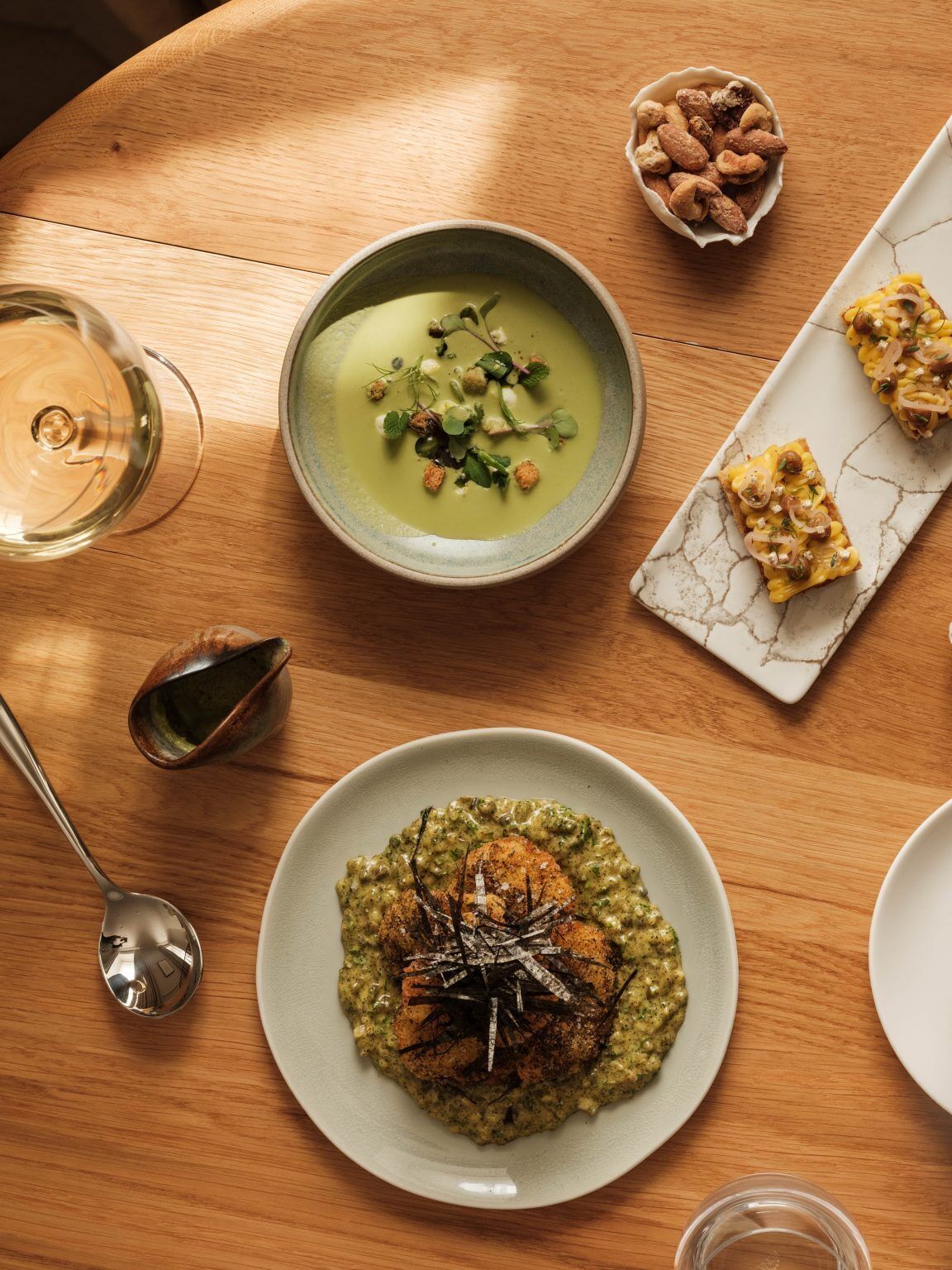 A dining table with a bowl of soup, a plate with a green dish, a small bowl of nuts, and a glass of wine.