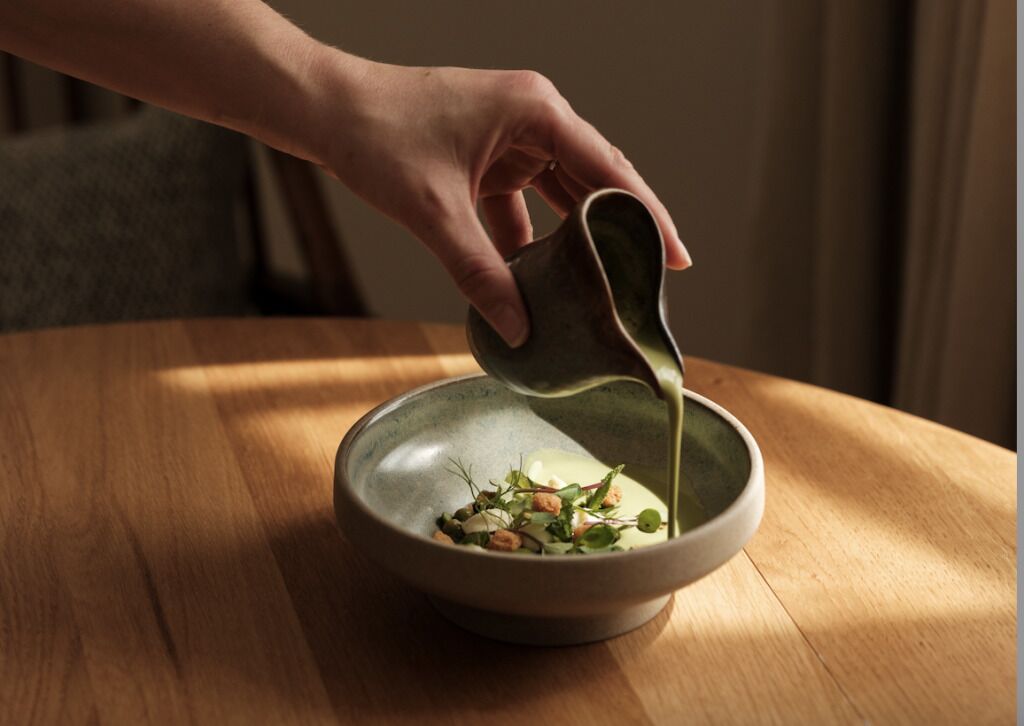 A hand pouring a liquid into a bowl with food on a wooden table.