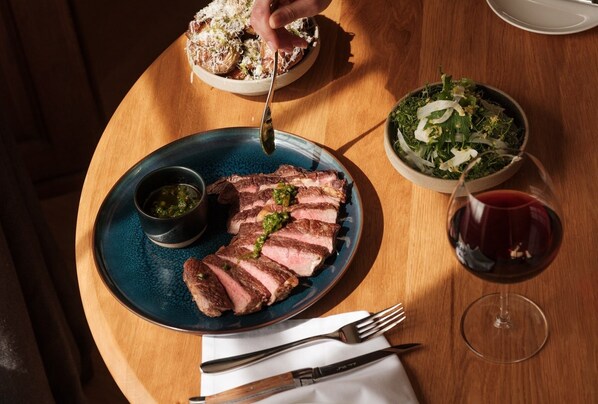 A plate of sliced steak with a green sauce, a bowl of salad, and a glass of red wine on a wooden table. - The Great Bustard (Salisbury)