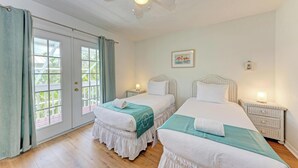 Une chambre avec deux lits, un ventilateur de plafond, une fenêtre avec des rideaux, une table de chevet avec une lampe et un tableau encadré au mur.