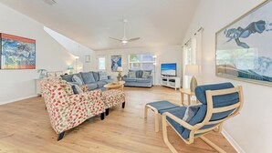 Un salon spacieux avec un plafond haut, un ventilateur de plafond et une grande fenêtre. On y trouve deux canapés, une table basse et une chaise en bois.