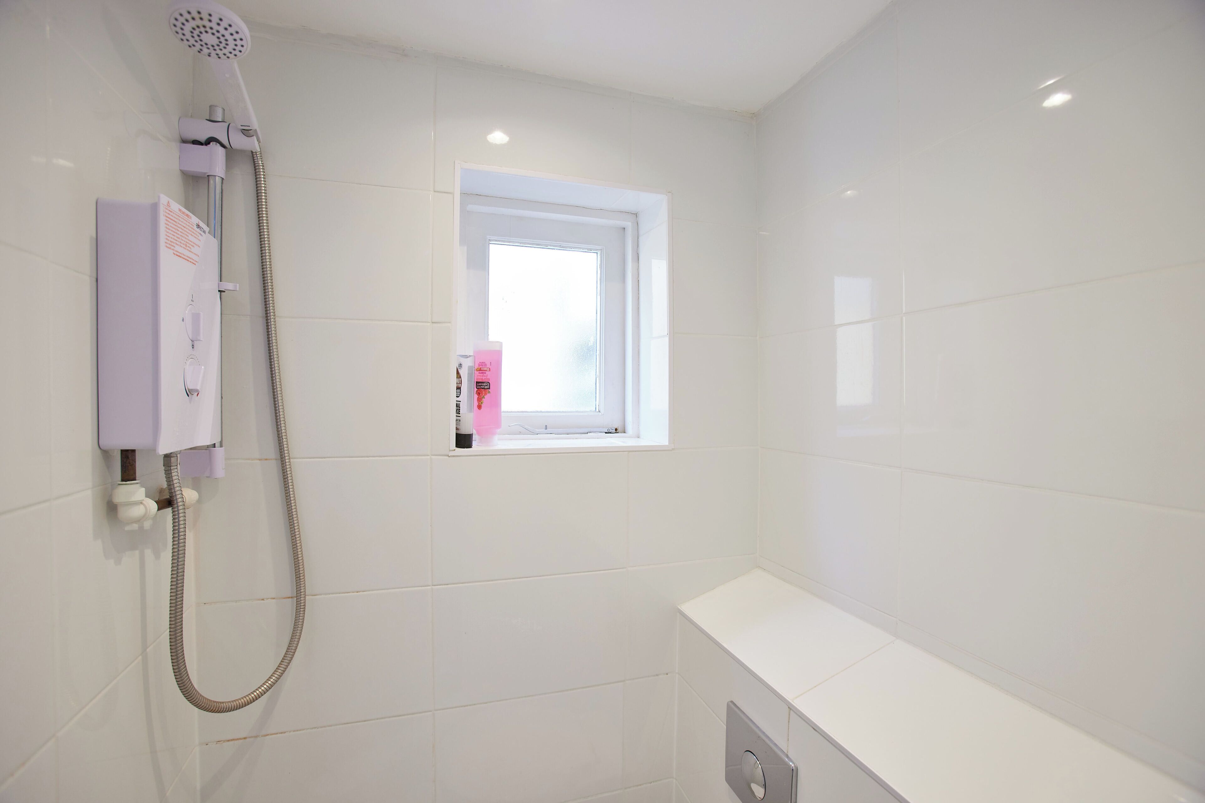 A small, white bathroom with a shower, a window, and a white sink.