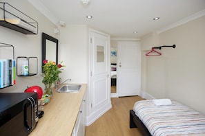 A compact bedroom with a bed, a bedside table, a mirror, a small kitchenette with a microwave, and a door leading to a bathroom.