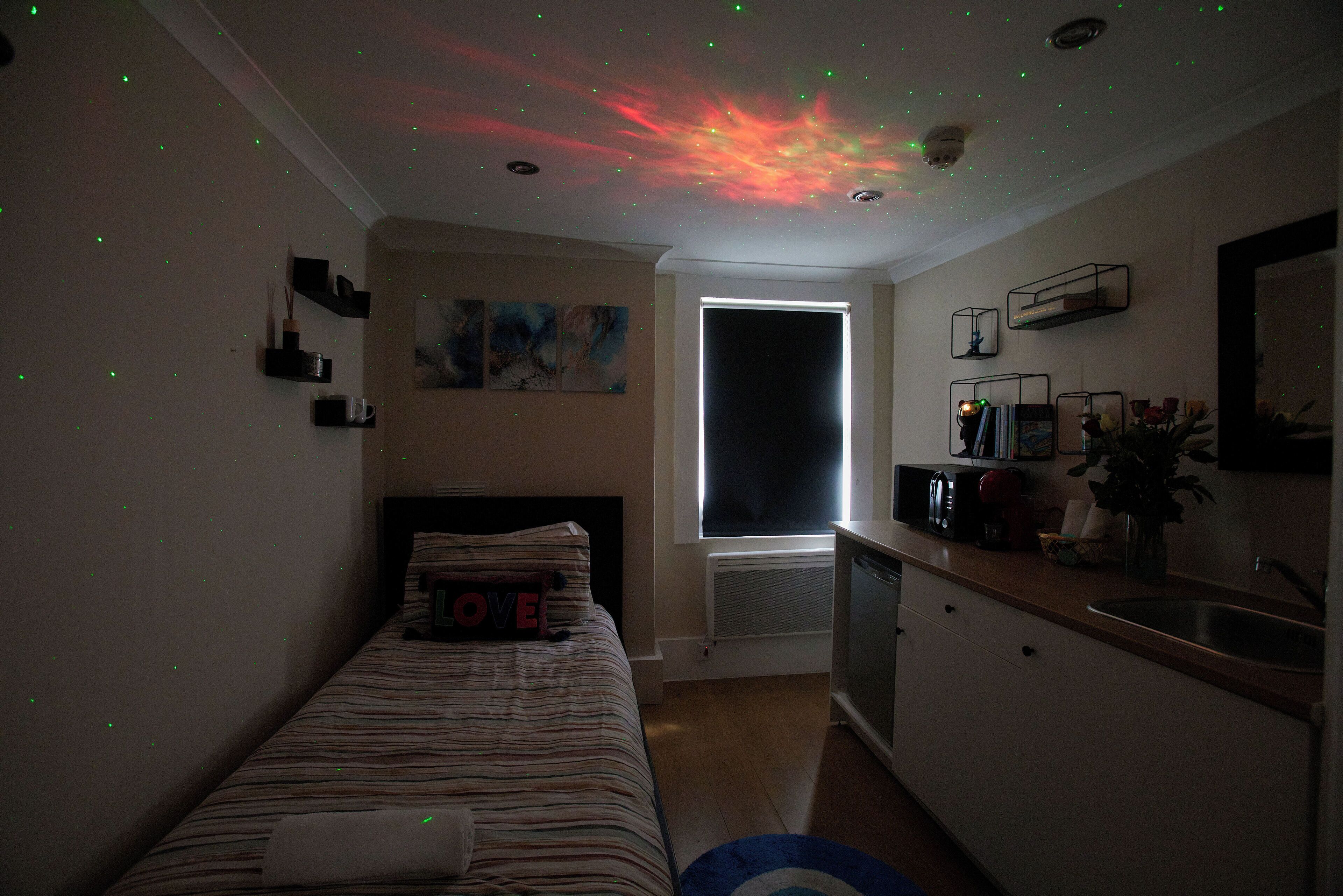 Ein Schlafzimmer mit Bett, Nachttisch, Kommode, einem Fenster und einem Deckenprojektor, der einen Sternenhimmel zeigt.