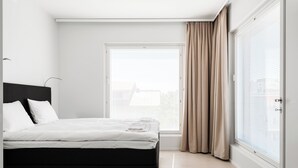 A modern bedroom with a black bed, white bedding, a large window with blinds, and a view of the city.