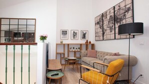 Una sala moderna con un sofá, un sillón amarillo, una mesa de centro y un televisor. Hay un mueble de madera con adornos y una obra de arte enmarcada en la pared.