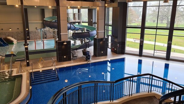A large indoor swimming pool with a spiral water slide, a staircase leading to a viewing area, and large windows offering a view of the outdoors.