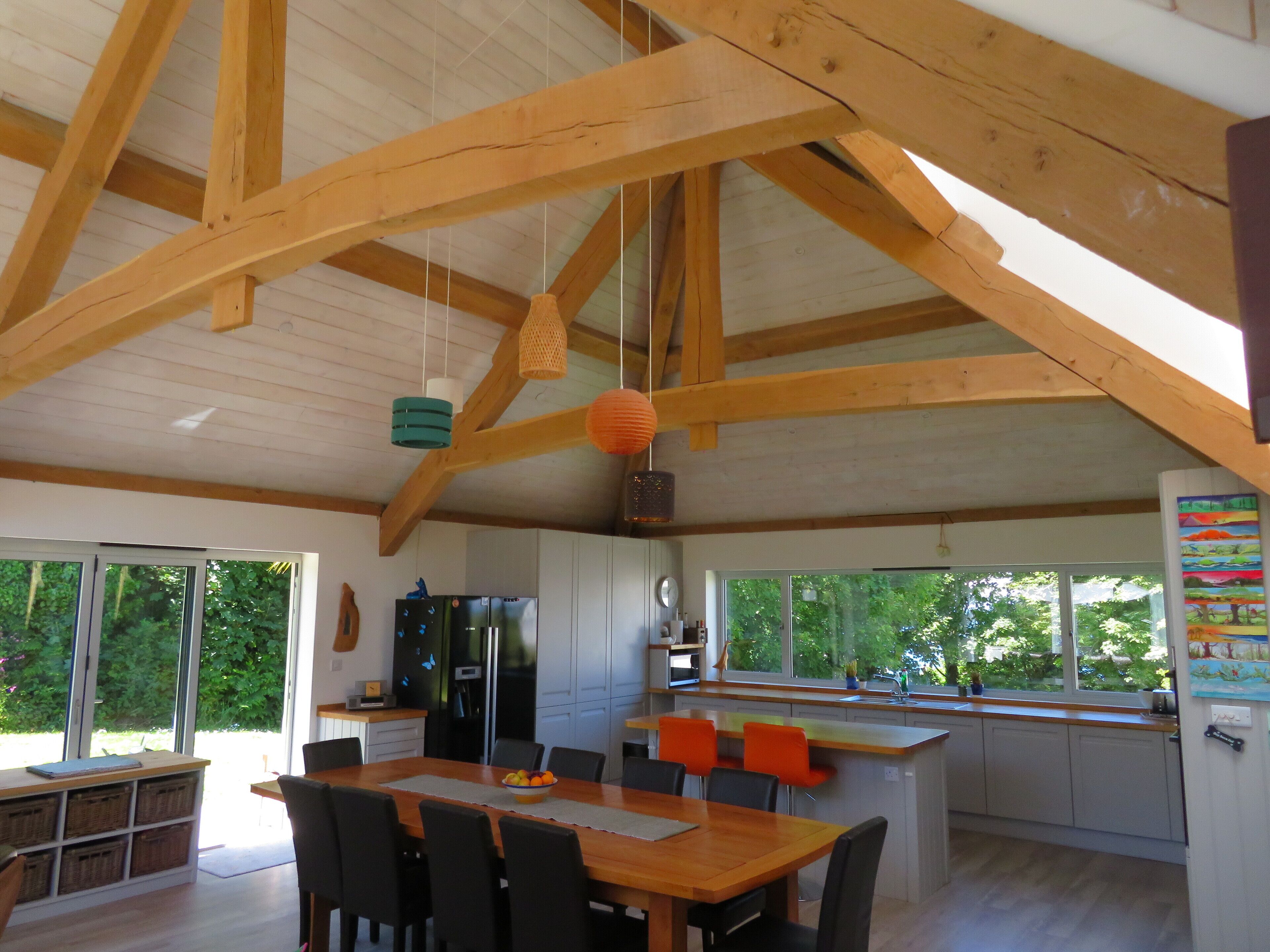 Eine geräumige Küche mit Holzbalken, eine Essecke mit Holztisch und -stühlen sowie große Fenster mit Blick auf Grünflächen.