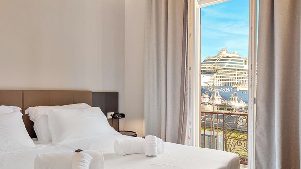 Een hotelkamer met een bed, witte lakens, een nachtkastje en uitzicht op een cruiseschip.
