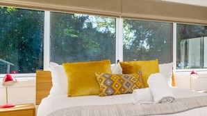 A neatly made bed with white linens and two mustard-colored pillows, a wooden headboard, and a view of trees through large windows. - Garden Precinct 3BR, Walking distance to Airport (Pialligo)