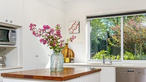 A kitchen with white cabinetry, a wooden countertop, a microwave, a dishwasher, a vase with pink flowers, and a window with a view of greenery. - Garden Precinct 3BR, Walking distance to Airport (Pialligo)