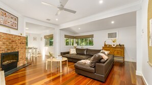 A spacious living room with a fireplace, a large leather sofa, wooden flooring, and a ceiling fan. - Garden Precinct 3BR, Walking distance to Airport (Pialligo)