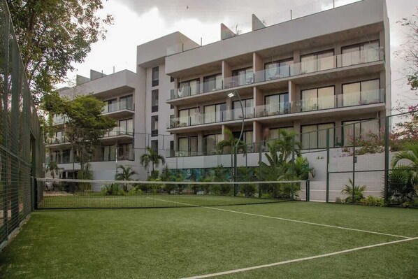 A multi-story residential building with balconies, a tennis court, and surrounding greenery.