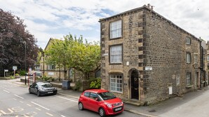 A stone building with a traditional architectural style, a clear street in front, and parked cars. - Grade II Quirky Yorkshire Cottage (ADDINGHAM)
