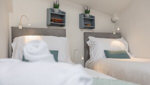 A hotel room with two beds, each with white linens and a teal pillow. There are wall-mounted shelves with decorative items and books. - Grade II Quirky Yorkshire Cottage (ADDINGHAM)