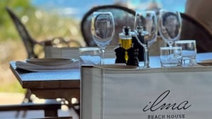 Ein Esstisch mit Meerblick, darauf eine Champagnerflasche und ein Glas Wasser. Der Tisch trägt die Aufschrift „ilma BEACH HOUSE“.