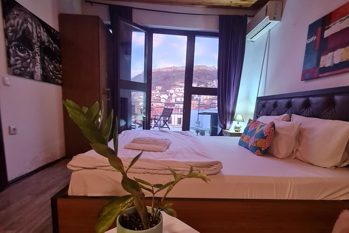Ein Schlafzimmer mit einem großen Bett, einer Topfpflanze und Blick auf eine Stadtlandschaft durch große Fenster.