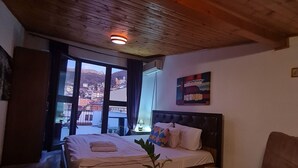 Camera d'albergo con soffitto in legno, un letto con vista sulla città, un tavolino con una pianta e una poltrona marrone.