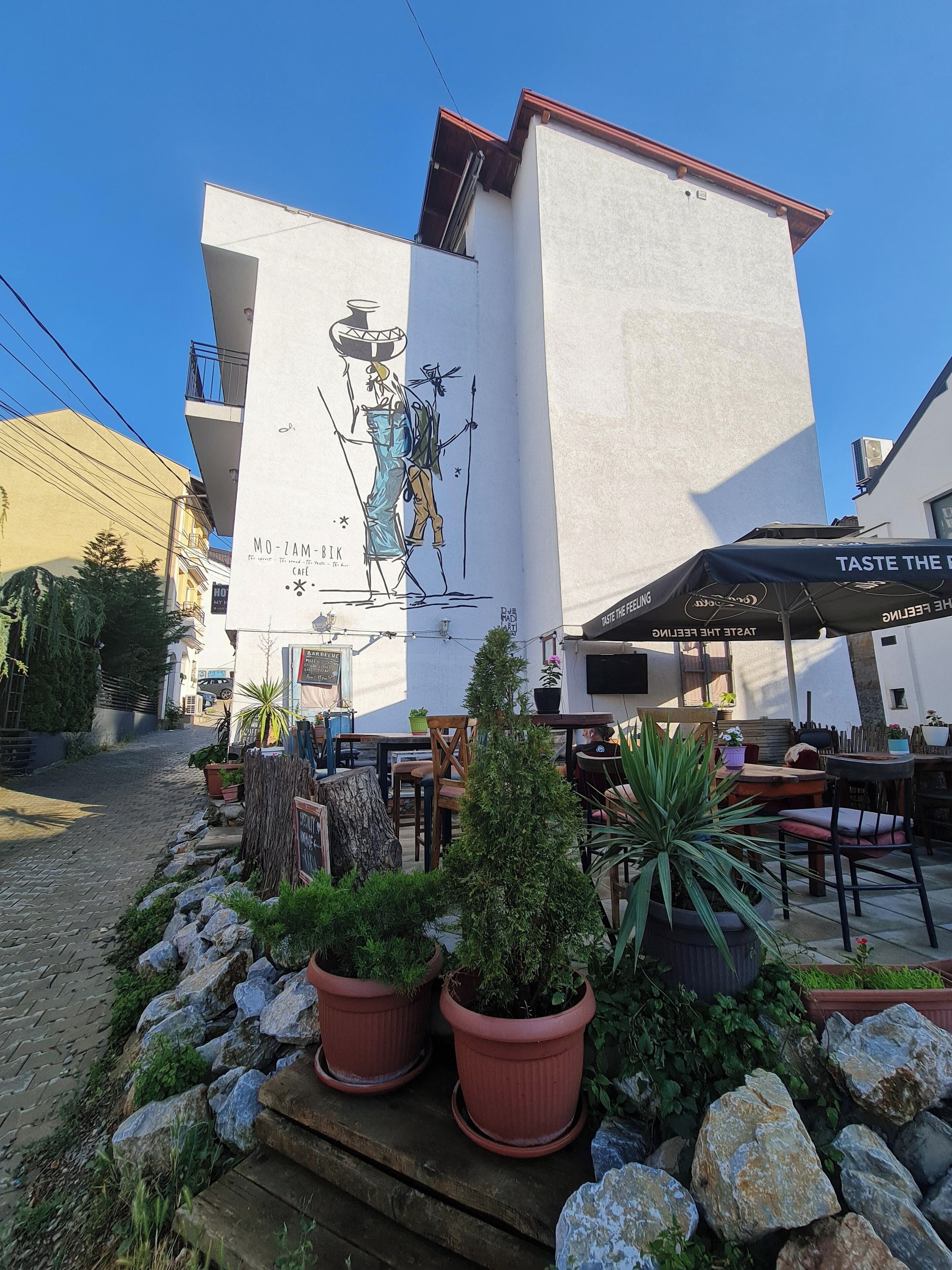 Un bâtiment orné d’une fresque représentant une figure vêtue d’un costume traditionnel. On y trouve des espaces de détente en plein air avec des tables et des chaises.