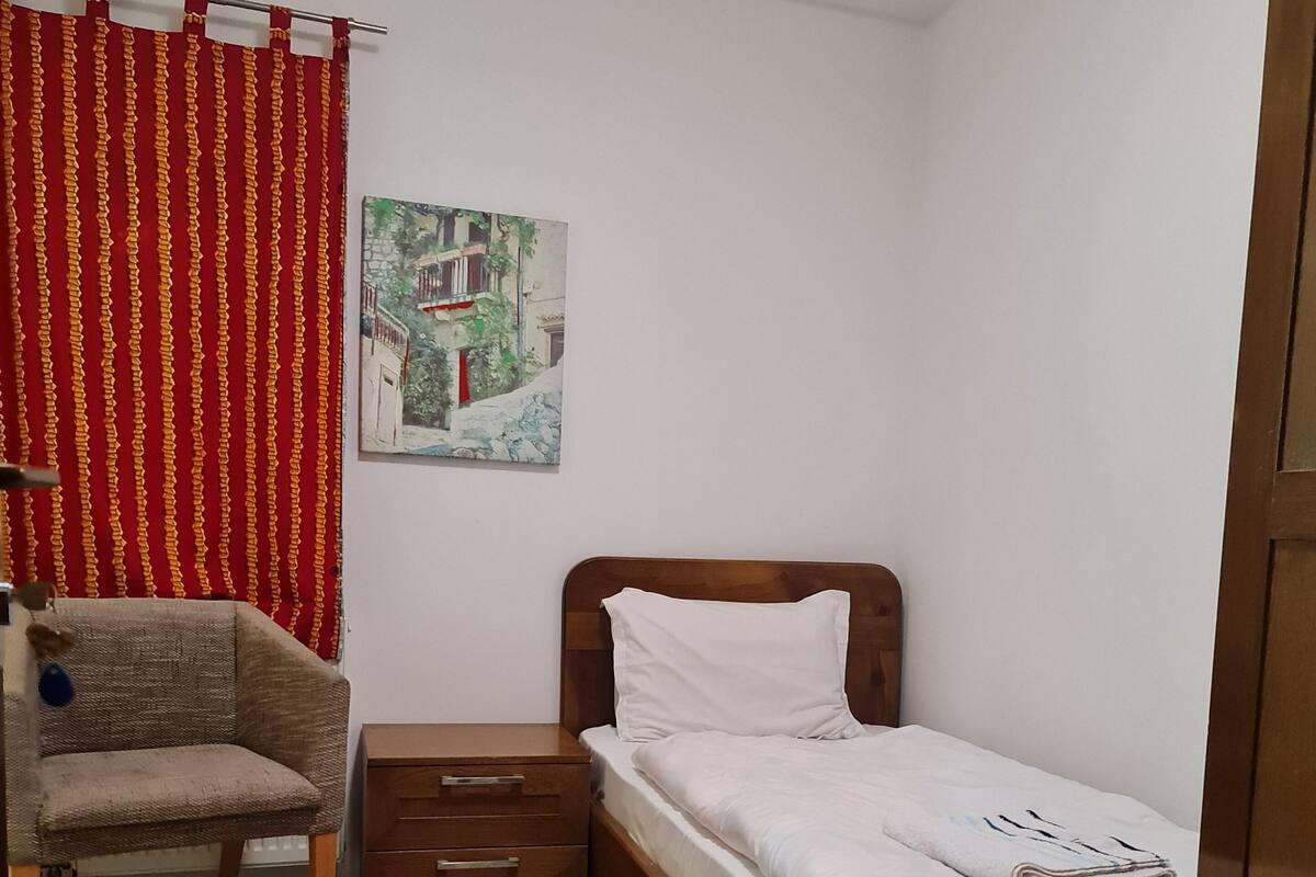 Ein Schlafzimmer mit einem hölzernen Bett, einem Sessel, einem kleinen Nachttisch aus Holz, einem roten Vorhang und einem Gemälde an der Wand.