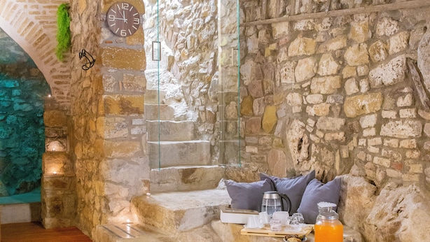 A stone-walled room with a wooden floor, a stone bench with cushions, a glass door, and a clock. - PORTA SOPRANA Luxury guest house &Spa (Sclafani Bagni)