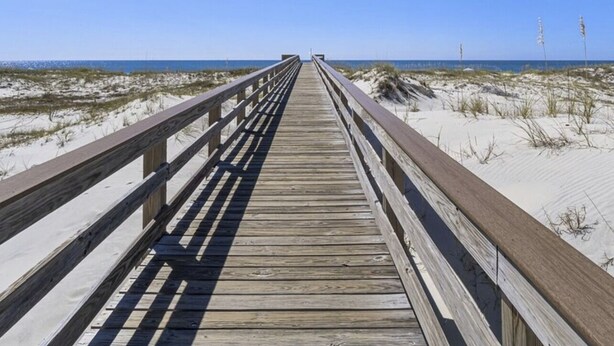 Ein Holzsteg führt zu einem Strand mit Dünen und einem klaren, blauen Himmel.