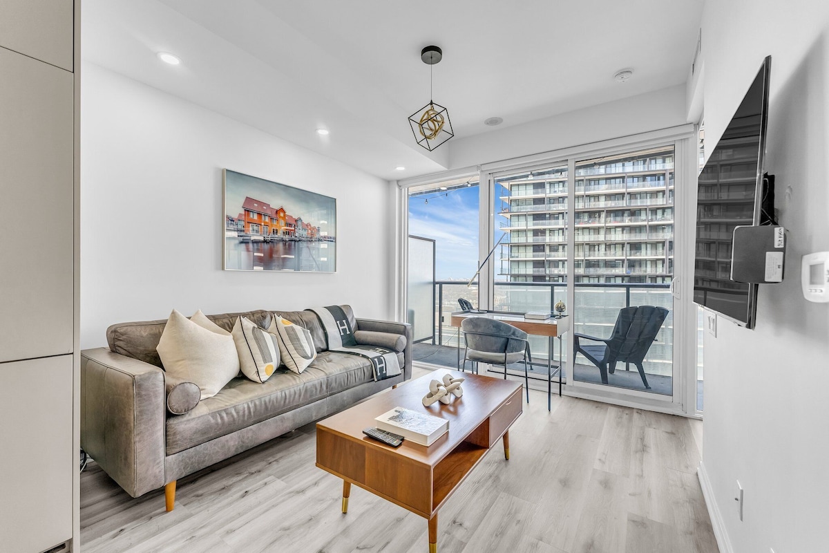 A modern living room with a grey sofa, a wooden coffee table, and a large window with a city view.