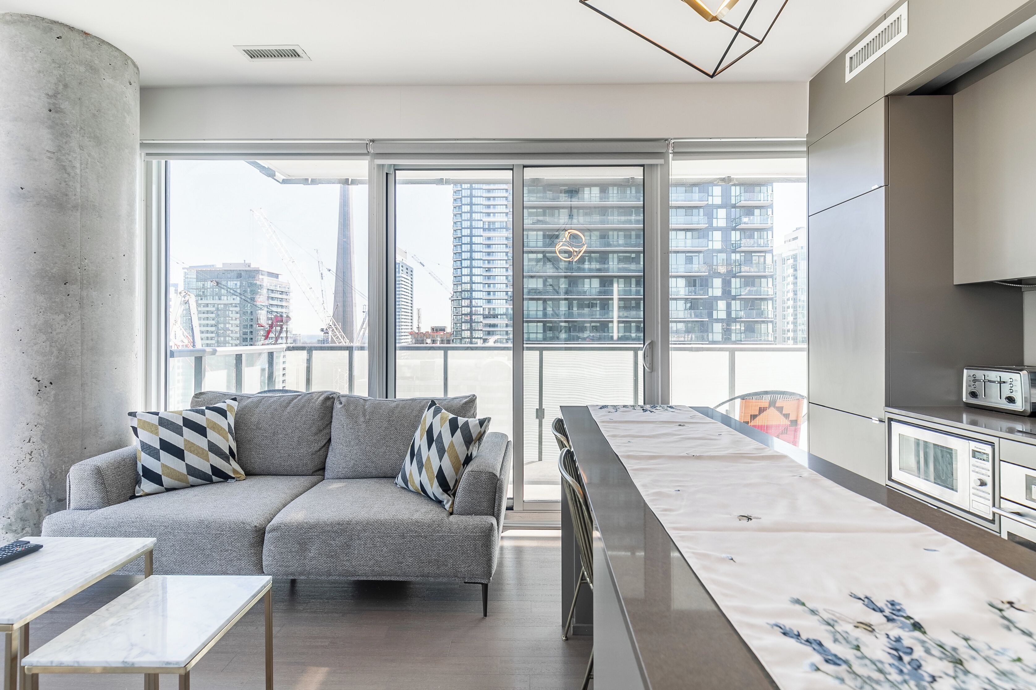 A modern apartment with a grey sofa, a marble coffee table, a kitchen with grey cabinets, and large windows offering a city view.