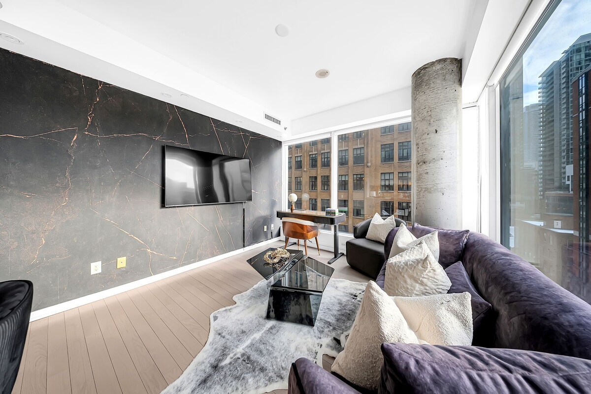 A modern living room with a large TV, a piano, and a view of a cityscape through large windows.
