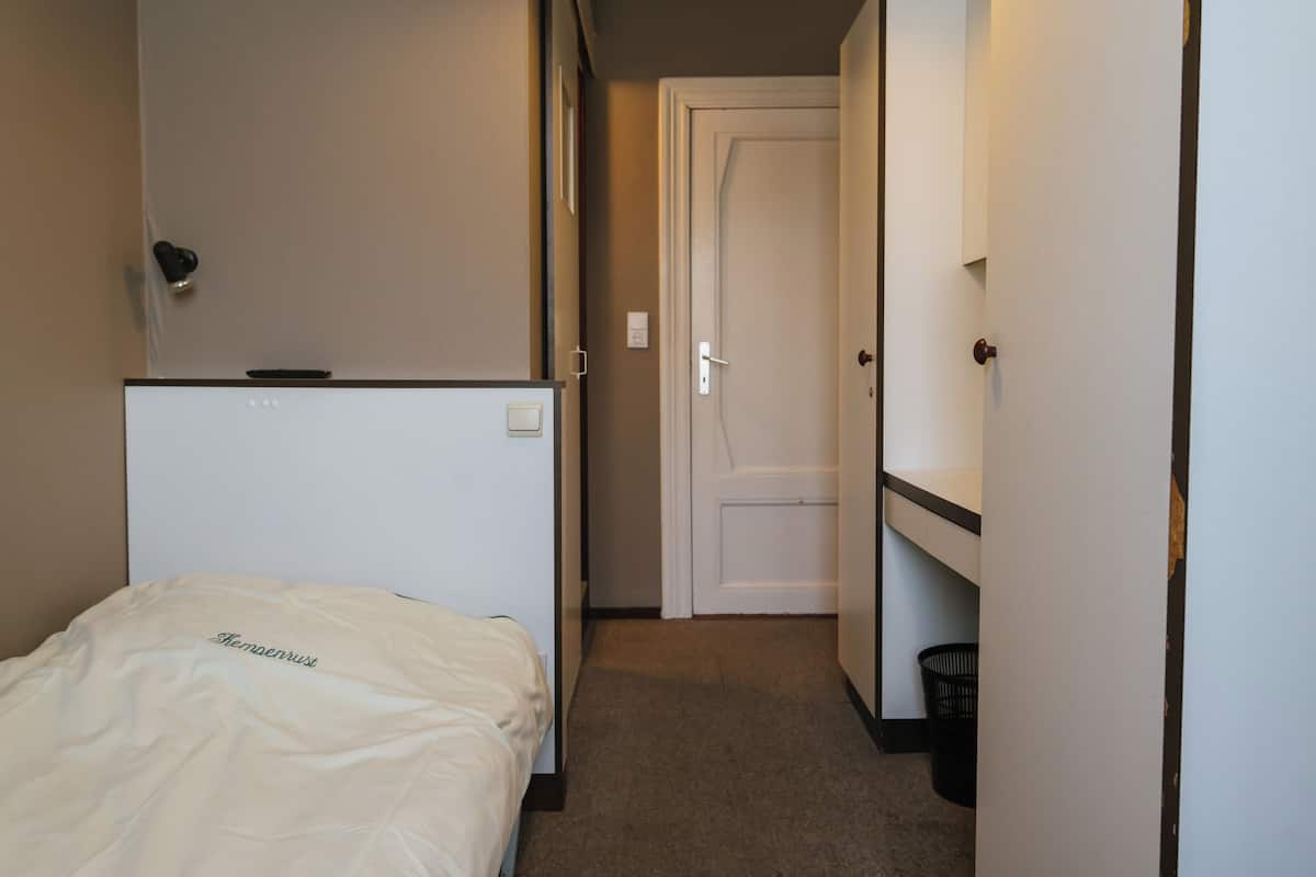Quarto de hotel com cama, porta, um cesto de lixo pequeno e luminária na parede.