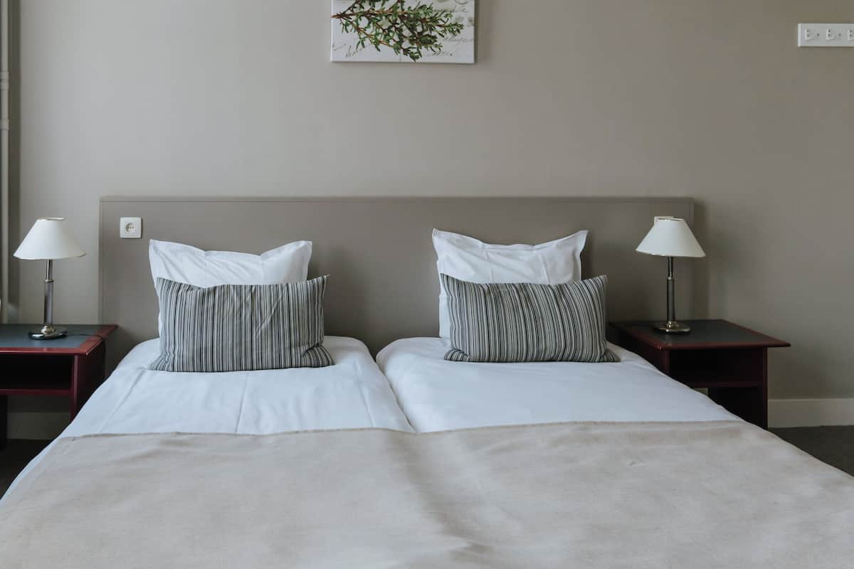 Quarto de hotel com duas camas, cada uma com edredom branco e almofadas listradas. Há mesas de cabeceira com luminárias e um interruptor de luz na parede.