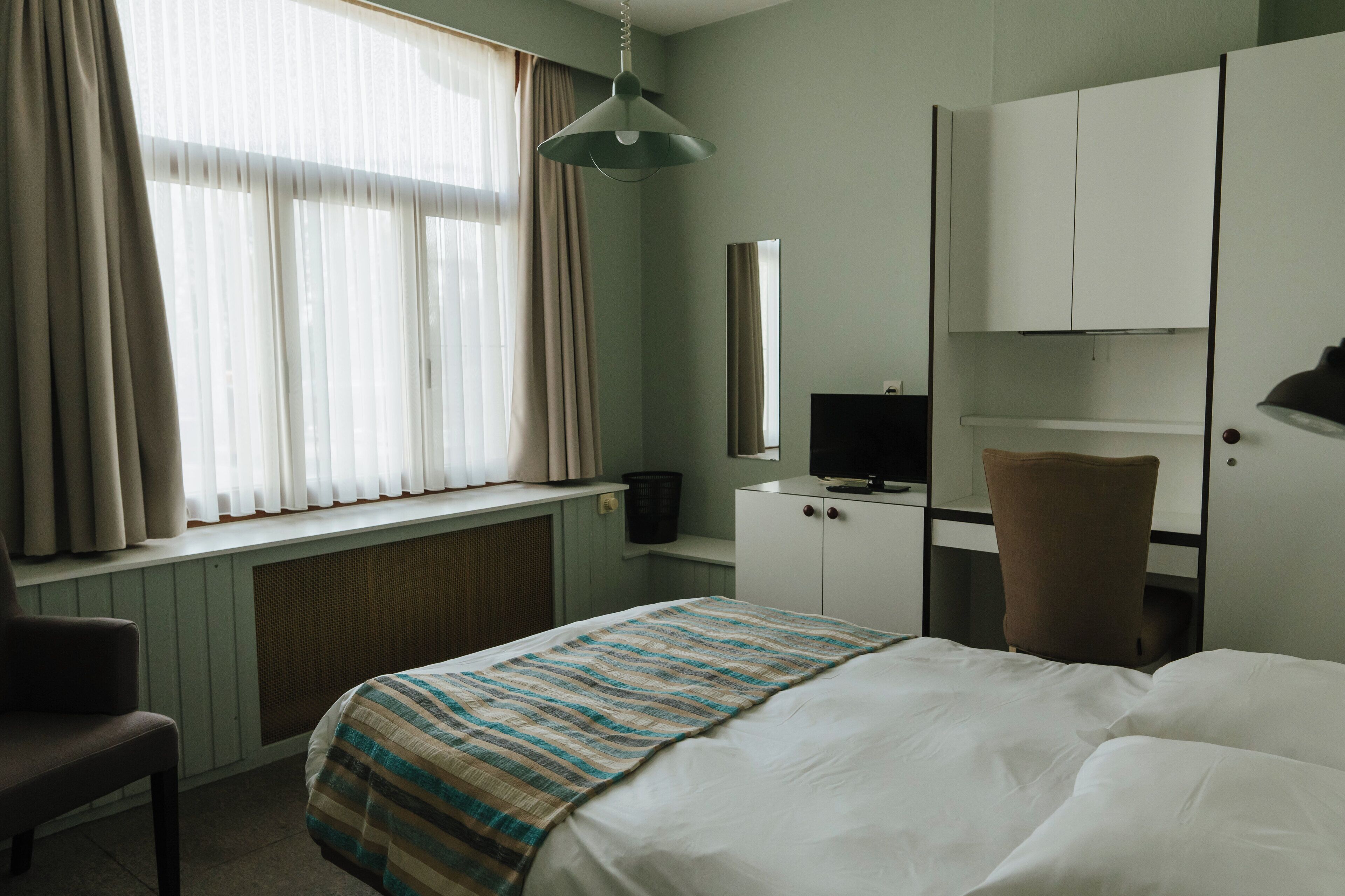 Een hotelkamer met een bed, een bureau, een stoel, een televisie, een kledingkast en een raam met gordijnen.