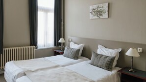 Una cama doble con sábanas blancas, dos mesitas de noche con lámparas, un radiador y una ventana con cortinas.