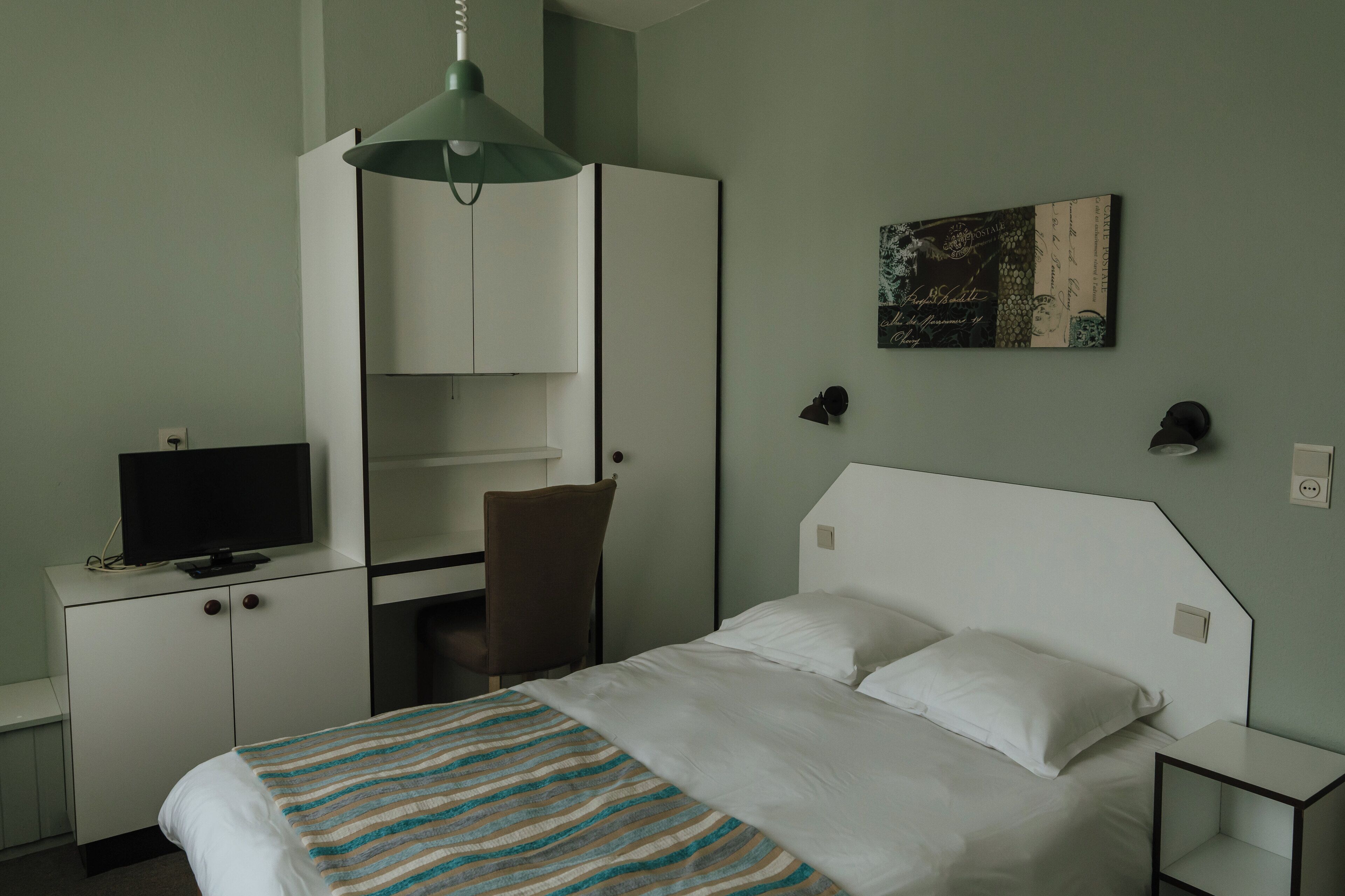 A hotel room with a bed, a desk with a television, a chair, and a wall-mounted shelf.