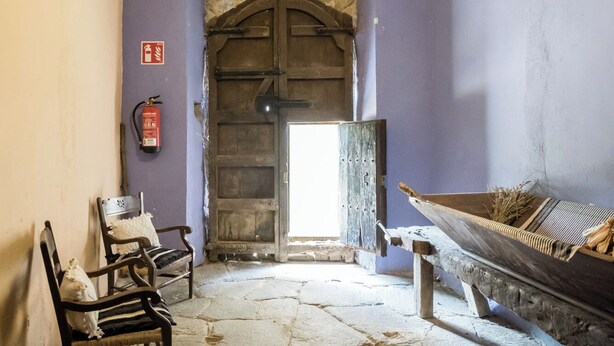 Una habitación rústica con piso de piedra, bancos de madera, una puerta grande de madera y una lámpara colgante.