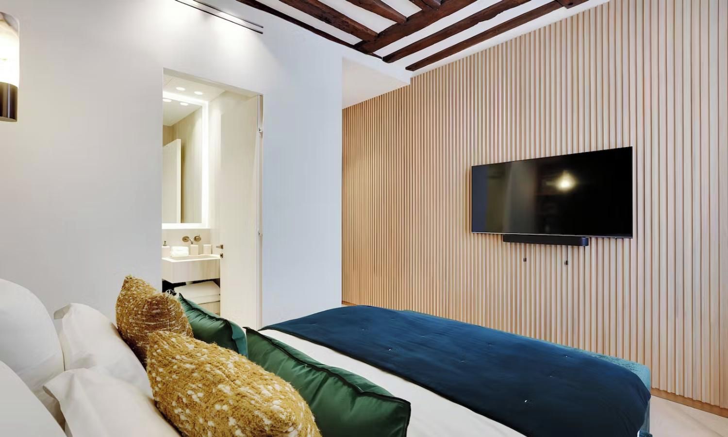Une chambre à coucher moderne, avec un lit, un téléviseur fixé au mur et une salle de bain visible à travers une porte ouverte.