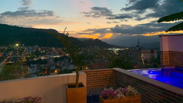 Una terraza en la azotea con piscina, vistas a un paisaje urbano al anochecer y una cadena montañosa al fondo.