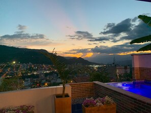 A rooftop terrace with a pool, a view of a cityscape at dusk, and a mountain range in the background. - Hotel Santorini (San Gil)
