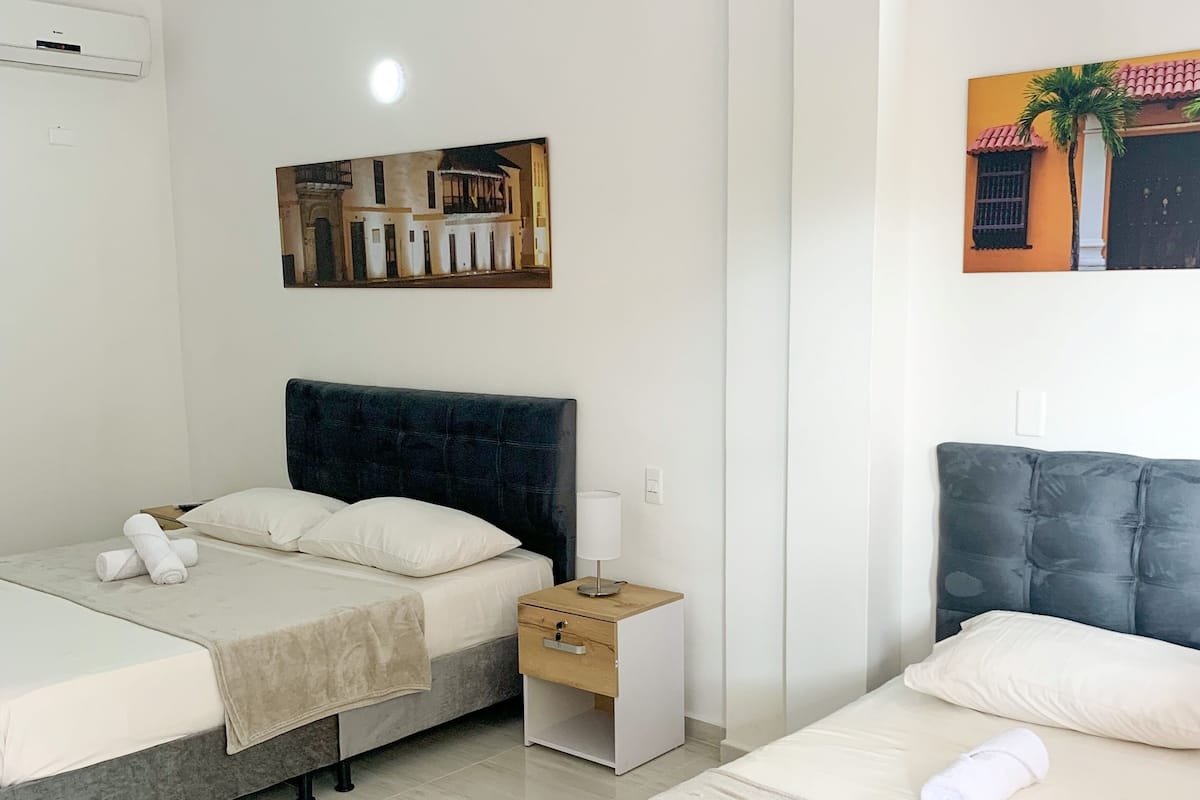 A hotel room with two beds, a nightstand with a lamp, a wall-mounted air conditioner, and a painting of a building.
