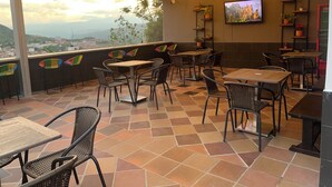 Un área exterior con mesas y sillas, un televisor montado en la pared y vistas de la ciudad.