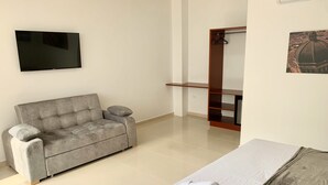 Una habitación de hotel moderna con un sofá gris, una cama con una manta gris, un televisor de pantalla plana montado en la pared y una estantería de madera.