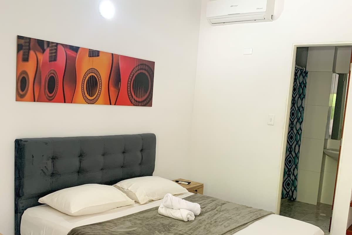 A modern bedroom with a large bed, a dark headboard, a wall-mounted air conditioner, and a colorful abstract painting above the bed.