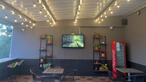 Un área de estar al aire libre con mesas y sillas, un televisor montado en la pared y estantes con plantas en macetas.