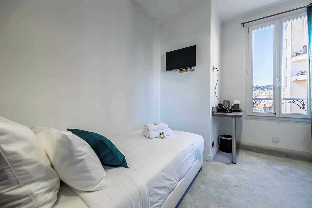 Una camera da letto con un letto, una TV fissata al muro, un comodino con un cestino e una finestra con vista sugli edifici.