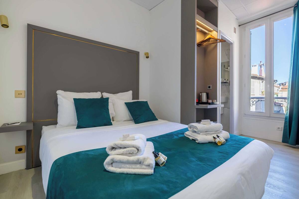 Camera d'albergo con un letto grande, biancheria da letto color acquamarina e vista sulla città attraverso un'ampia finestra.