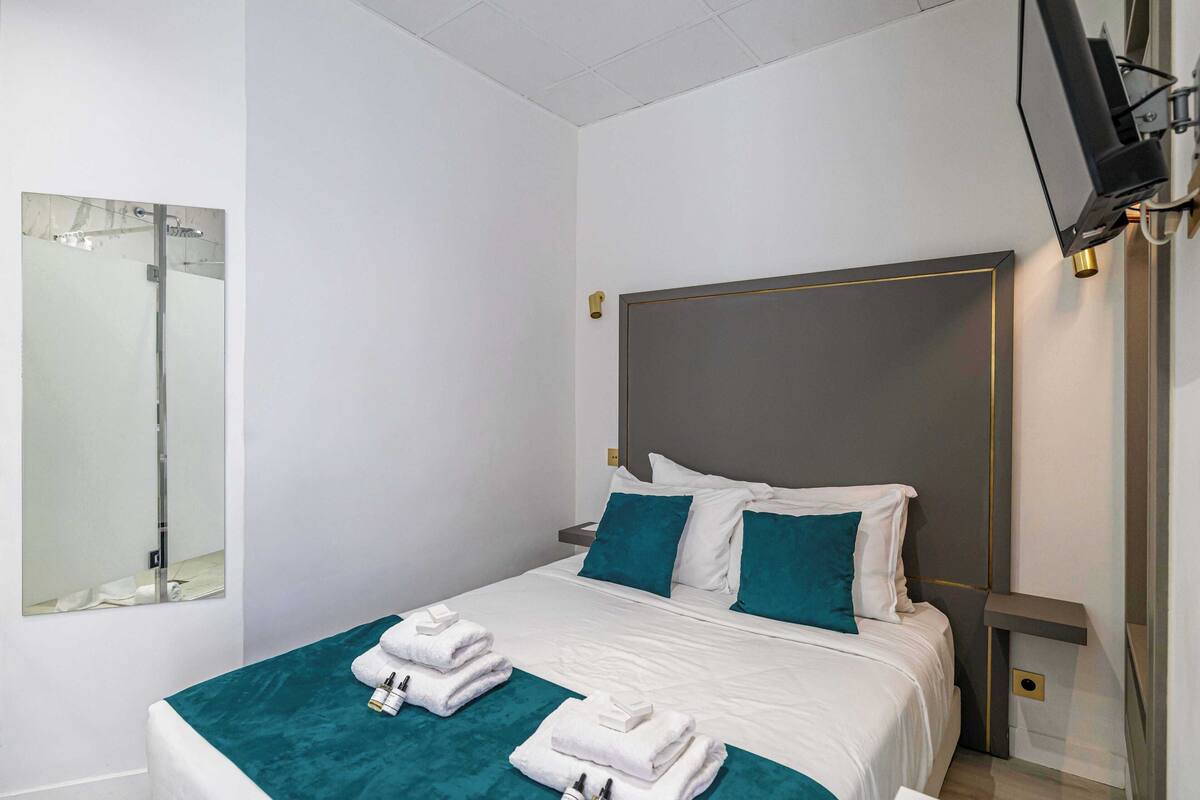 Una camera d'albergo moderna con un letto grande, copripiumino color petrolio, lenzuola bianche, uno specchio ampio e un televisore su un mobile.