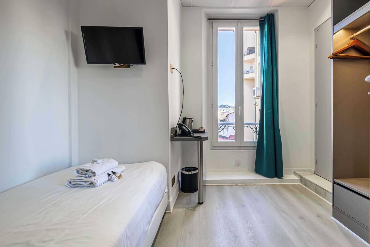 Una camera d'albergo con un letto, una televisione fissata al muro, una finestra con tende e un tavolino con un cestino.