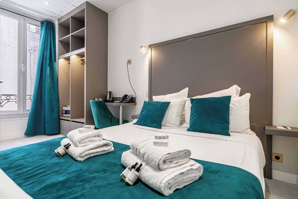 Una camera d'albergo moderna con un letto rifatto, biancheria da letto color petrolio, un comodino con il telefono e un armadio a muro.