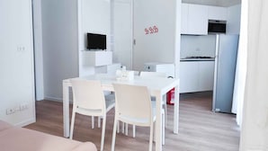 A modern, minimalist kitchen with white cabinetry, a built-in oven, and a microwave. There is a dining area with a white table and chairs, and a flat-screen TV mounted above the table. - Holiday apartment near the sea (Jesolo)