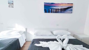 A hotel room with two single beds, a large bed with a black mattress, white pillows, and a painting of a sunset over the ocean. - Holiday apartment near the sea (Jesolo)